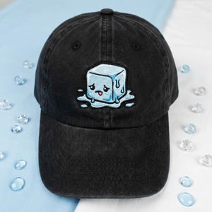 May include: Black baseball cap with a distressed finish. The cap features an embroidered design of a cartoon ice cube with a sad expression and melting water droplets. The ice cube is light blue, and the cap has a curved brim.