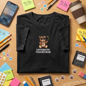May include: Black t-shirt with a cartoon bear wearing sunglasses and holding a coffee cup and a magic wand. The text "CAFFEINATED TEACHER BEAR" is below the bear graphic. The shirt is surrounded by school supplies.