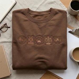 May include: A brown sweatshirt featuring embossed medical imaging symbols: radiation, lead vest, MRI, X-ray, and a monitor. The crew neck sweatshirt is folded on a neutral surface, with coffee and a notebook in the background.