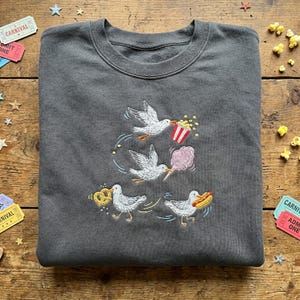 May include: A charcoal grey crewneck sweatshirt featuring an embroidered design of four white seagulls. One seagull carries a red and white striped popcorn bucket, another holds pink candy floss, one carries a pretzel, and one holds a hot dog.