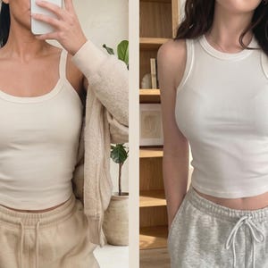 May include: Two white ribbed tank tops shown in a side-by-side comparison. The left tank top features wide shoulder straps and a scoop neckline. The right tank top features a high crew neckline and racerback-style armholes. Both are fitted.