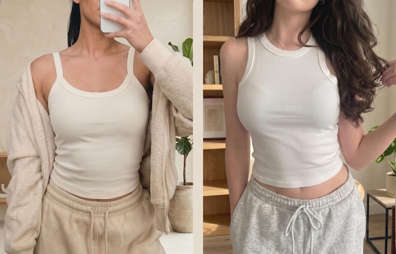 May include: Two white ribbed tank tops shown in a side-by-side comparison. The left tank top features wide shoulder straps and a scoop neckline. The right tank top features a high crew neckline and racerback-style armholes. Both are fitted.