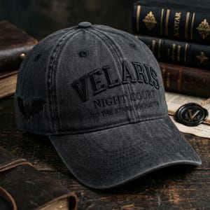 May include: A dark grey baseball cap featuring the word "VELARIS" arched across the front, with "NIGHT COURT" and "TO THE STARS WHO LISTEN" below. A bat graphic is on the side. The cap is made of a worn, textured fabric.