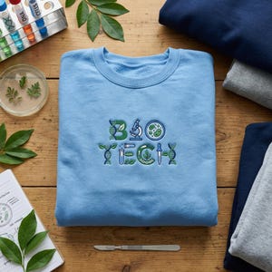 May include: A light blue sweatshirt with the embroidered text "BIO TECH" in green and blue. The design incorporates science-themed graphics like DNA strands, a microscope, and a petri dish. The sweatshirt is folded on a wooden surface.