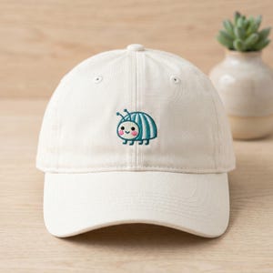 Peut inclure: Casquette de baseball blanche avec une broderie d'insecte de dessin animé. L'insecte a un corps rayé bleu et blanc, des antennes et un visage souriant. La casquette a une visière incurvée et un bouton sur le dessus.