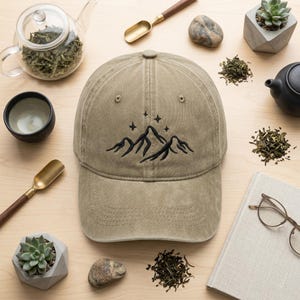 May include: A khaki baseball cap featuring a black embroidered mountain and star design. The image includes tea-related items such as a teapot, teacups, and loose tea leaves. The cap is made of a soft, worn material.
