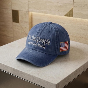 Könnte beinhalten: Blaue Denim-Baseballkappe mit weißer Stickerei "We the People serve no king". Auf der Seite befindet sich ein Aufnäher mit der amerikanischen Flagge. Die Kappe liegt auf einer weißen Oberfläche.