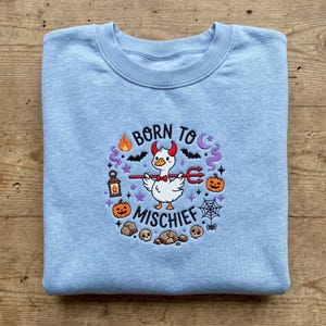 May include: A light blue crewneck sweatshirt featuring an embroidered design of a white duck with red devil horns holding a red trident. The text BORN TO MISCHIEF surrounds the duck, accompanied by pumpkins, bats, a lantern, and a spider web.
