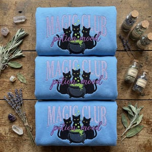 May include: Three light blue sweatshirts with the embroidered text "MAGIC CLUB potion mood" and a graphic of black cats around a bubbling cauldron. The sweatshirts are displayed on a wooden surface with dried herbs and small glass jars.