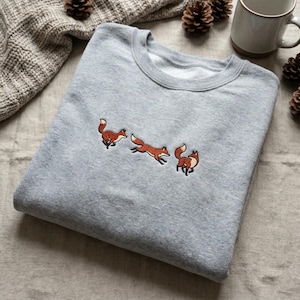 Könnte beinhalten: Ein hellgraues Sweatshirt mit gestickten Füchsen in einer Laufsequenz. Die Füchse sind in Orange-, Braun- und Weißtönen mit schwarzen Umrissen dargestellt. Das Sweatshirt hat einen Rundhalsausschnitt und lange Ärmel, ideal für Freizeitkleidung.