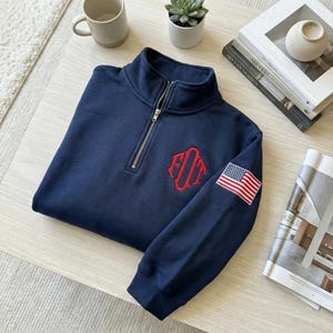 May include: Navy blue quarter-zip sweatshirt with a red embroidered monogram and an American flag patch on the sleeve. The sweatshirt has a silver zipper and is folded on a light-colored surface.