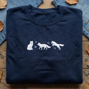 May include: Navy blue sweatshirt featuring a white embroidered design of three foxes and butterflies. The foxes are depicted in different poses, with one sitting, one walking, and one jumping. The design is centered on the chest of the sweatshirt.