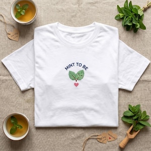 May include: A white short-sleeved t-shirt featuring an embroidered design in the center. The design includes two green mint leaves with a small smiling face, a pink heart below, and the dark blue text MINT TO BE arched above the leaves.