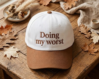 May include: A white baseball cap with a brown brim and a brown button on top. The front of the cap features the embroidered brown text "Doing my worst" in a serif font. The cap has a structured crown with ventilation eyelets.