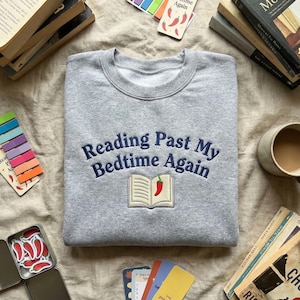 Könnte beinhalten: Ein hellgraues Sweatshirt mit dem aufgestickten Schriftzug "Reading Past My Bedtime Again" in Marineblau. Das Design zeigt ein offenes Buch mit einem roten Chili. Bücher und Lesezubehör umgeben das Sweatshirt.