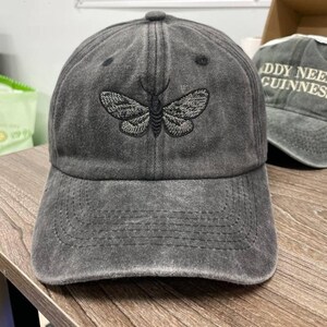 May include: A gray baseball cap with a distressed finish. The cap features an embroidered moth design on the front. The cap has a curved brim and an adjustable strap at the back. Another cap with text is visible in the background.