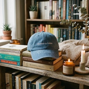 May include: A blue denim baseball cap with the embroidered text "enemies to lovers" sits atop a stack of books. The image is set in a cozy, book-filled environment with a lit candle and a scented candle.