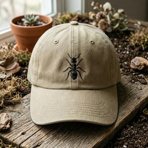 May include: A beige baseball cap featuring a black embroidered ant. The cap has a curved peak and a six-panel design. The hat is displayed on a wooden surface.