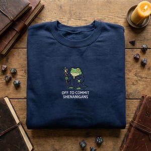 May include: Navy blue sweatshirt featuring an embroidered frog wizard design with a staff. The text "OFF TO COMMIT SHENANIGANS" is below the frog. The sweatshirt is on a wooden surface with books, dice, and a candle.