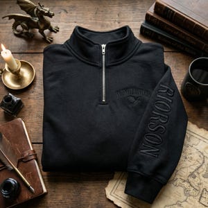 May include: Black quarter-zip sweatshirt with the word "WINGLEADER" embroidered on the chest and "RIORSON" on the sleeve. The sweatshirt has a high collar and a silver zipper. The design includes a bird graphic. The sweatshirt is folded on a wooden surface.