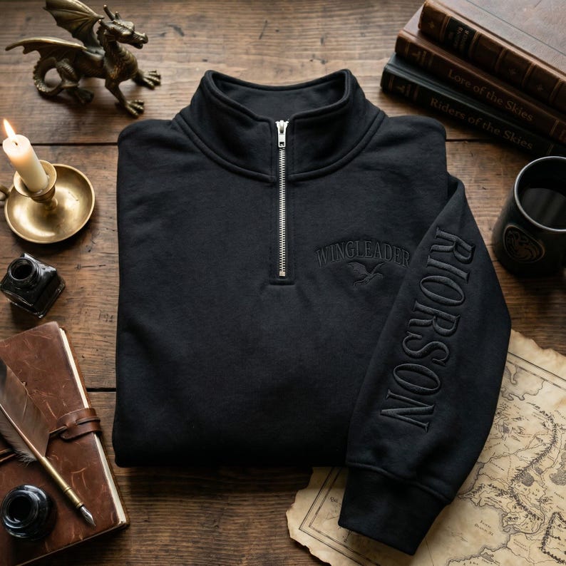 May include: Black quarter-zip sweatshirt with the word "WINGLEADER" embroidered on the chest and "RIORSON" on the sleeve. The sweatshirt has a high collar and a silver zipper. The design includes a bird graphic. The sweatshirt is folded on a wooden surface.