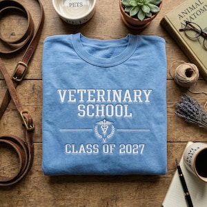 May include: A light blue sweatshirt with white embroidered text reading "VETERINARY SCHOOL CLASS OF 2027" and a caduceus symbol. The sweatshirt is folded on a wooden surface, with a leather leash, a pet bowl, and a coffee cup nearby.