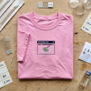 May include: A light pink t-shirt featuring a central embroidered graphic of a computer window labelled MICROBE.EXE NOT RESPONDING. The graphic depicts a green microbe sleeping inside a petri dish with small zzz symbols above it.
