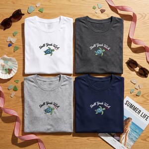 May include: Four t-shirts in white, charcoal, grey, and navy blue, each embroidered with a turtle and the text "Shell Yeah USA." Sunglasses, sea glass, and a magazine with the words "Summer Life" are also visible.