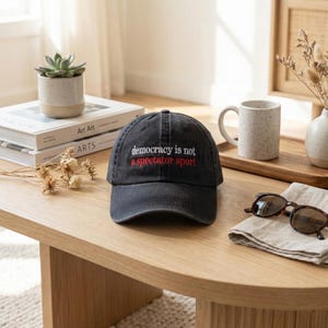 May include: A dark grey baseball cap with the embroidered text "democracy is not a spectator sport" in white and red. The cap is on a light wood table, with other items in the background.