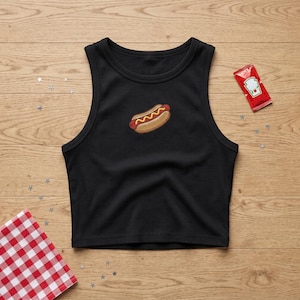 May include: A black ribbed crop top tank shirt featuring a small embroidered hot dog graphic in the center. The design includes a brown bun with a red sausage and yellow mustard. A small red ketchup packet is positioned to the upper right.
