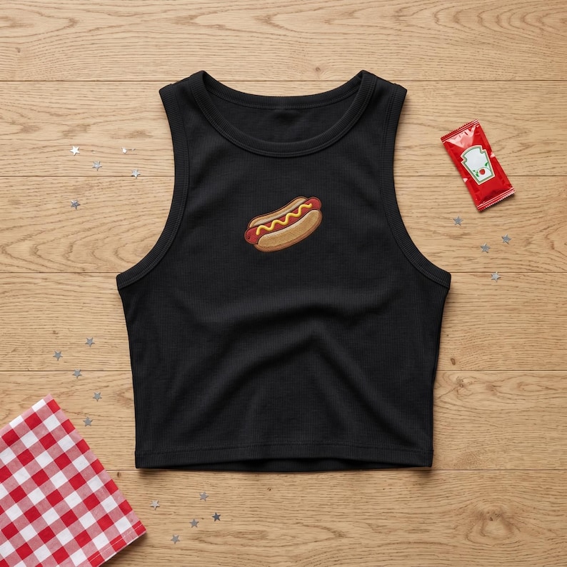 May include: A black ribbed crop top tank shirt featuring a small embroidered hot dog graphic in the center. The design includes a brown bun with a red sausage and yellow mustard. A small red ketchup packet is positioned to the upper right.
