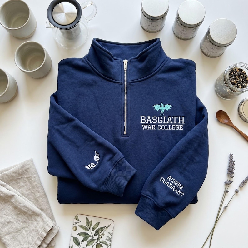 May include: Navy blue quarter-zip sweatshirt with the text "BASGIATH WAR COLLEGE" and a dragon graphic. The left sleeve has a white wing design, and the right sleeve reads "RIDERS QUADRANT". The sweatshirt is surrounded by tea-related items.