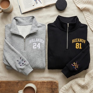 May include: Two quarter-zip sweatshirts, one gray and one black, with hockey-themed embroidery. The gray sweatshirt features "HOLLANDER 24" on the chest and crossed hockey sticks on the sleeve. The black sweatshirt has "ROZANOV 81" and similar sleeve embroidery.