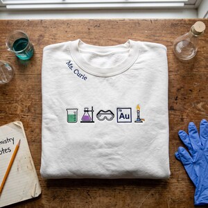 May include: A white crewneck sweatshirt featuring embroidered science-themed icons including a green beaker, a purple flask, safety goggles, the gold periodic table symbol Au, and a Bunsen burner, with the text Ms. Curie embroidered near the collar.