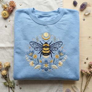 May include: A pale blue sweatshirt with an embroidered bee design. The bee is surrounded by flowers, a crescent moon, and stars. The bee has black and yellow stripes. The flowers are blue and yellow. The sweatshirt is folded.