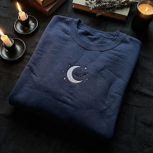 May include: Navy blue sweatshirt with an embroidered crescent moon and bat design. The moon is silver, and the bat is black. The sweatshirt is a crew neck style and is folded neatly. The design is centered on the chest.