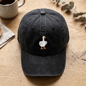 May include: A black baseball cap with a washed-out, vintage-style finish. The front center of the cap features a small, embroidered graphic of a white goose with an orange beak and orange feet, standing in a forward-facing position.