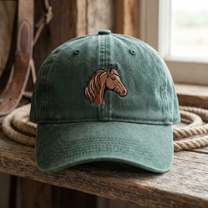 May include: A green baseball cap featuring a brown horse head embroidery. The cap is made of soft fabric and has a curved peak. The hat is displayed on a wooden surface.