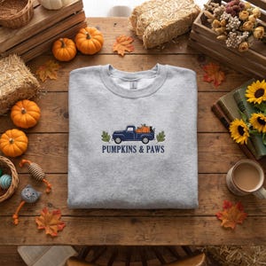 Peut inclure: Un sweat-shirt gris clair à col rond avec une broderie représentant un pick-up bleu foncé transportant des citrouilles orange et un chat noir. Le texte PUMPKINS & PAWS est brodé en bleu foncé sous le camion, encadré par deux feuilles vertes.