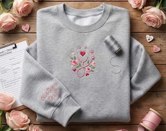 以下が含まれることがあります： ピンクの花とハートで刺繍された聴診器のデザインが施された、ヘザーグレーのスウェットシャツ。袖には「custom text」の文字がピンクで刺繍されています。ピンクのバラと医療メモがスウェットシャツの周りに配置されています。