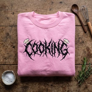 May include: A light pink crewneck sweatshirt featuring black embroidered text that reads COOKING in a jagged, thorny font style. The design includes two small white chef hats placed at the beginning and end of the word, centered on the chest.