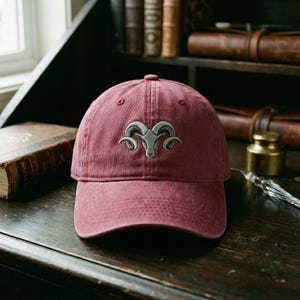 May include: A maroon baseball cap with a curved peak. The cap has a grey embroidered ram's head logo. The hat is made of a soft, textured fabric and has a classic six-panel design.