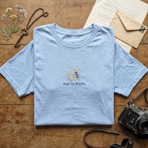 May include: A light blue t-shirt featuring an embroidered gold locket design with blue glasses inside. Below the locket, the text "kept in drafts" is embroidered in dark blue thread. The shirt has a crew neckline and short sleeves.