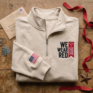 May include: A beige quarter-zip sweatshirt featuring a black embroidered text that reads WE WEAR RED next to a red banner with white stars and the text REMEMBER EVERYONE DEPLOYED. A small American flag patch is on the left sleeve cuff.