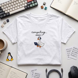May include: A white t-shirt featuring a graphic of a white goose wearing black headphones and using a dark blue laptop. Above the goose is the text "compiling..." with a loading bar graphic. The design is centred on the chest of the shirt.