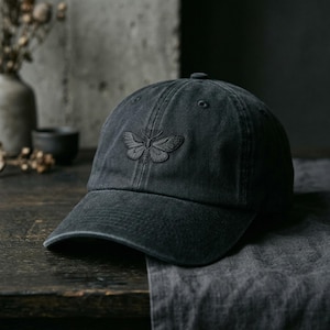 May include: A black baseball cap with a butterfly design. The cap is made of a soft, textured fabric and features a raised butterfly emblem on the front. The cap has a curved brim and an adjustable strap.