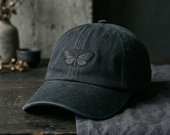 May include: A charcoal gray baseball cap featuring a tonal embroidered moth design on the front center panel. The cap has a structured crown, a curved brim, and stitched eyelets for ventilation, crafted from a washed cotton fabric material.