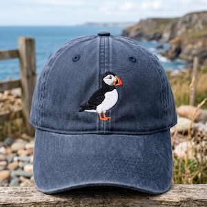 May include: A navy blue baseball cap featuring an embroidered puffin. The puffin has a white chest, black back, and orange beak and feet. The cap is made of a soft material and has a curved brim. The background shows a coastal scene.