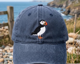 May include: A navy blue baseball cap featuring an embroidered puffin. The puffin has a white chest, black back, and orange beak and feet. The cap is made of a soft material and has a curved brim. The background shows a coastal scene.