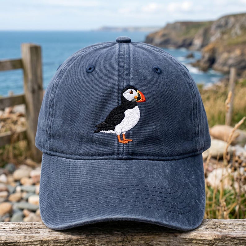 May include: A navy blue baseball cap featuring an embroidered puffin. The puffin has a white chest, black back, and orange beak and feet. The cap is made of a soft material and has a curved brim. The background shows a coastal scene.